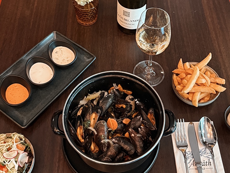 Geserveerde pan met verse mosselen in een zwarte pan, drie sausjes, friet, salade en een glas witte wijn (La Guirlande Sauvignon Blanc) op een donkerhouten tafel bij Amrâth Hotels.