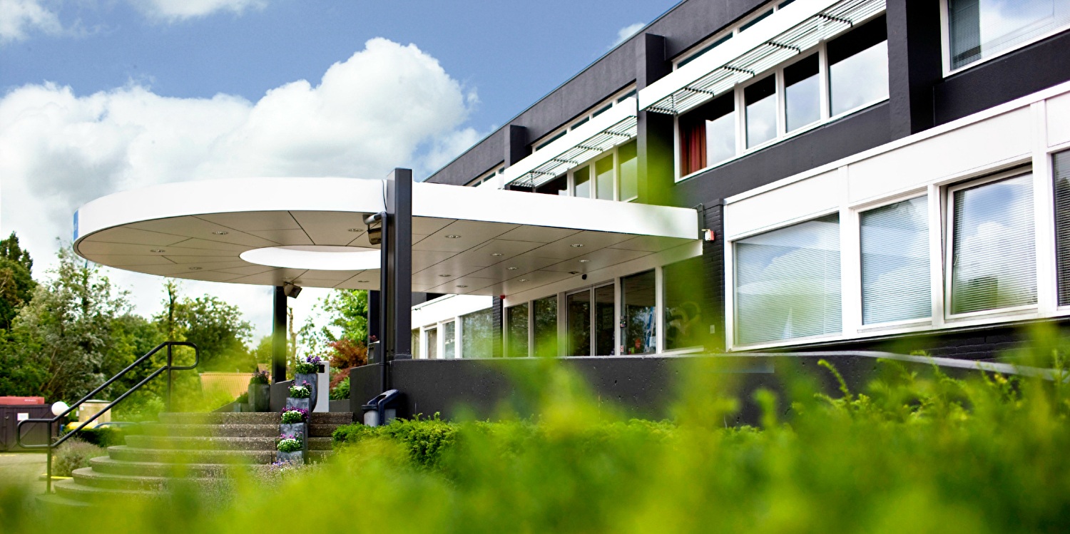 Ansicht der Vorderseite des Amrâth Rotterdam Airport Hotel mit modernem Eingang, Blumenkästen und umgebendem Grün unter teilweise bewölktem Himmel.