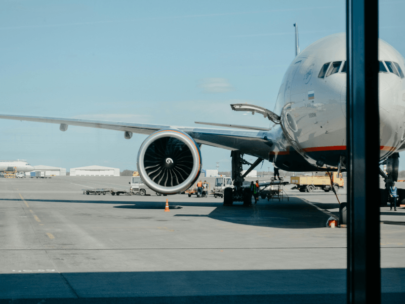 Groot passagiersvliegtuig op een luchthavenplatform gezien door een raam, met zicht op de motor, landingsgestel en grondpersoneel dat bezig is met laad- en voorbereidende werkzaamheden.