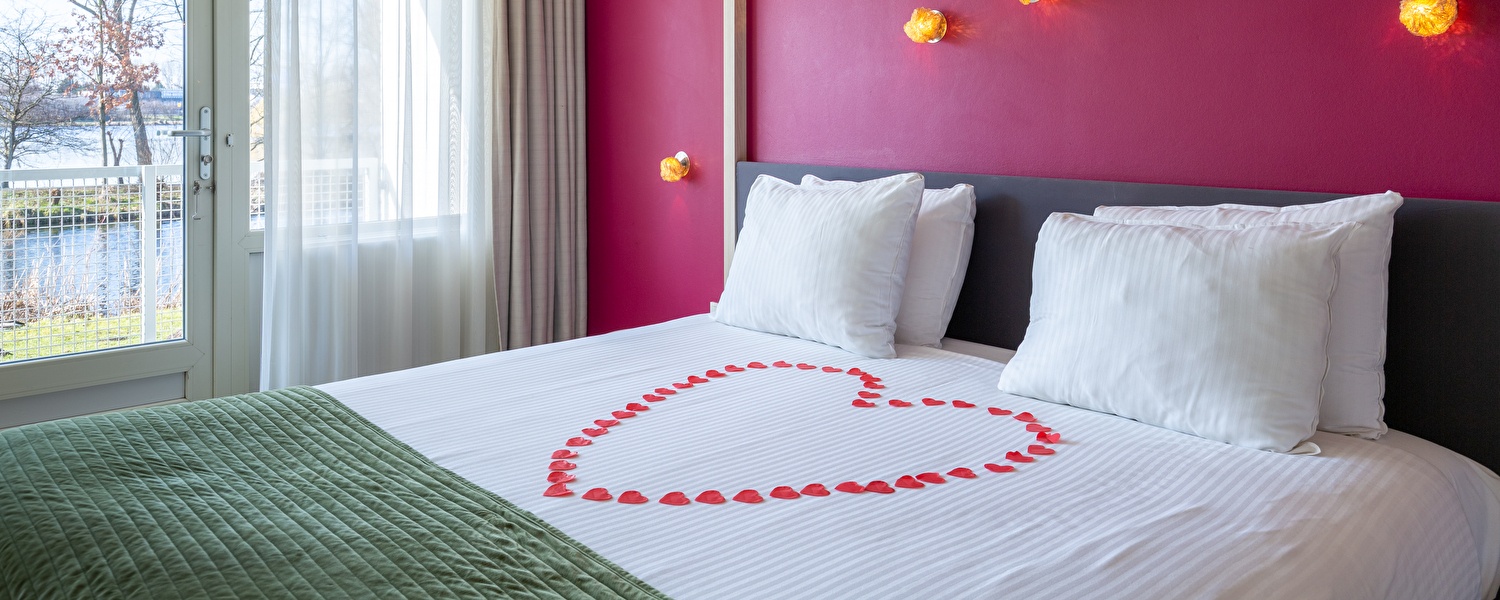 Romantically made bed with rose petals in heart shape in a hotel room at Amrâth Airport Hotel Rotterdam, with a view of the water.