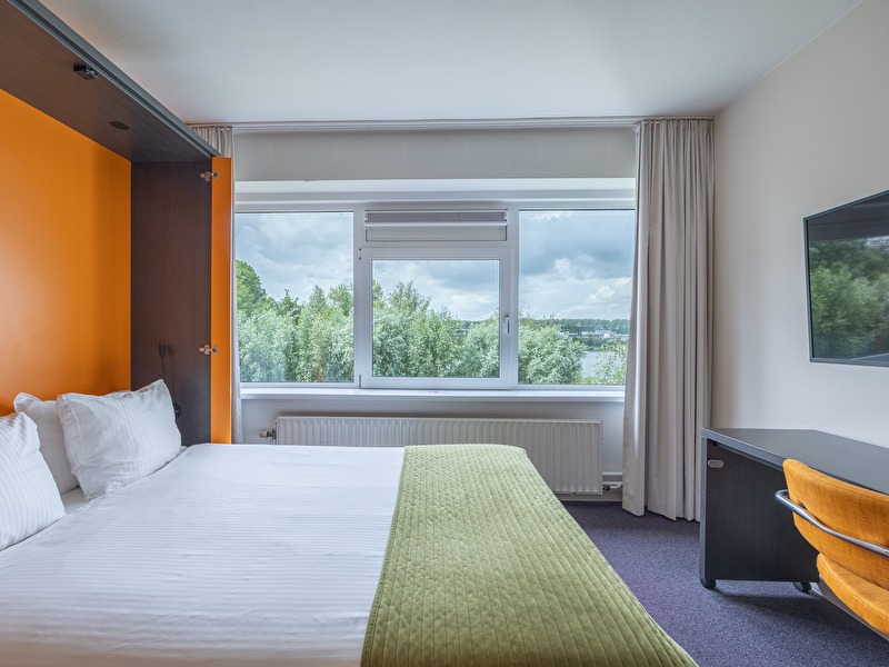 Hotelzimmer im Amrâth Rotterdam Airport Hotel, leuchtend orangefarbene Akzentwand, Schreibtisch mit Stuhl und Blick auf Bäume und Wasser durch ein großes Fenster.
