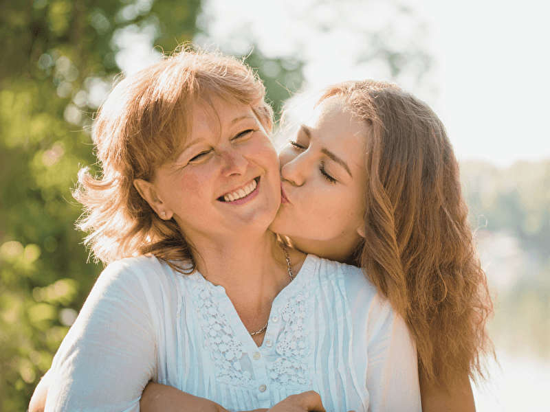 Laughing mother is lovingly embraced and kissed on the cheek by her daughter during a sunny day in a green, natural environment.